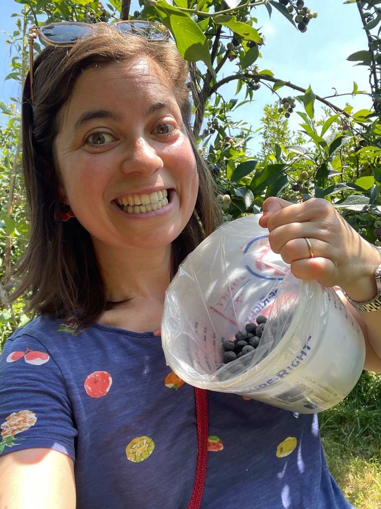 A white woman with brown hair smiles enthusiastically while holding a bucket of blueberries. 